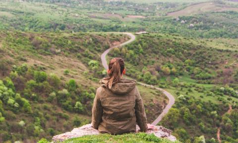 Woman sitting with her back to the camera on top of a rock overlooking treetops.