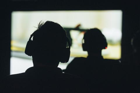 The silhouette of two people seen from the back, each wearing headphones and looking at a projected image on a movie screen. 