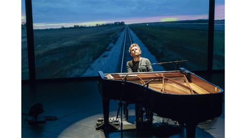A man sits at a black grand piano, his head tilted back, a spotlight illuminating him in a circle of light. Behind him is an image of an empty road stretching off to a sunset horizon.