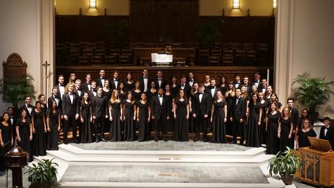 50 men and women stand in three rows on risers so they are all visible. The men wear black suits and the women wear long black dresses. Behind and above them is visible a large sheet of music which seems to be for the organist.